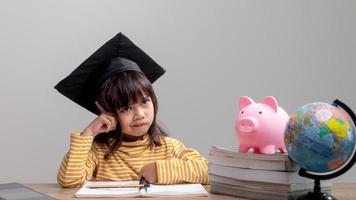 Asian little girl wearing a graduation cap with a pink piggy bank, Saving money, investment the future, photo