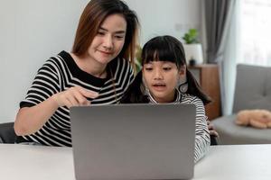 Asian little young girl kid learning online class at home with mother. Preschool child use laptop computer do homework, homeschool from school teacher by digital remote internet with support from mom. photo