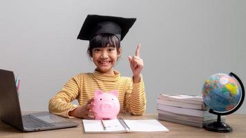 niña asiática con una gorra de graduación con una alcancía rosa, ahorrando dinero, invirtiendo en el futuro, foto