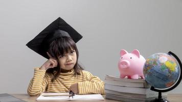 Asian little girl wearing a graduation cap with a pink piggy bank, Saving money, investment the future, photo