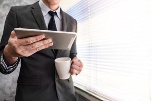 Close up of businessman using tablet and holding coffee cup while standing at a window in an office photo