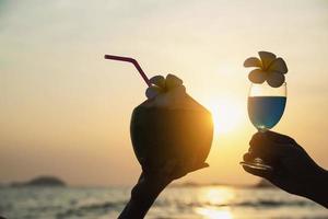 Silhouette couple hand holding cocktail glass and fresh coconut decorated by plumeria flower on beach background - happy couple relax honeymoon celebration vacation with sea nature concept photo