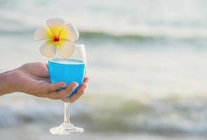 Closeup woman hand holding cocktail glass decoration with plumeria flower with beach background - happy relax celebration vacation in sea nature concept photo