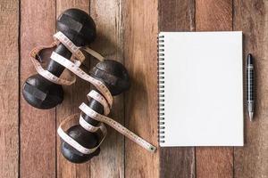 Top view of iron dumbbells with measure tape and notebook on wooden table background. photo