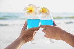 Pareja asiática sosteniendo una decoración de copa de cóctel con flor de plumeria con fondo de playa de mar de olas - feliz celebración de vacaciones de relax en el concepto de naturaleza marina foto