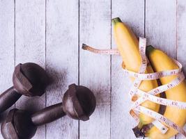 plátanos con cinta métrica y pesas de hierro en el fondo de la mesa de madera. concepto de entrenamiento y dieta foto