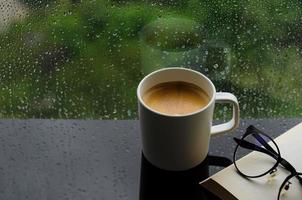 A cup of hot coffee with book and spectacles on table in morning with rain drop on window and green background. Stay home and relaxing concept. photo