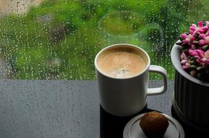 A cup of hot coffee with small cake on table in morning with rain drop on window and green background. Stay home and relaxing concept. photo