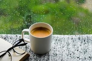 A cup of hot coffee with book and spectacles on table in morning with rain drop on window and green background. Stay home and relaxing concept. photo