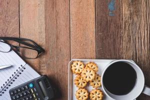 Top view of Notebook with glasses,cookie and coffee cup on wooden table background. Free space for text photo