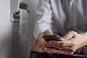 Man sitting on toilet bowl while playing mobile phone - health problem concept photo