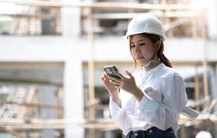 Architect with a blueprints at a construction site. Portrait of woman constructor wearing white helmet and safety yellow vest. Upset sad, skeptical, serious woman looking at the phone screen outdoors. photo
