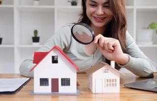 Happy woman hand holding magnifying glass and looking at house model, house selection, real estate concept. photo