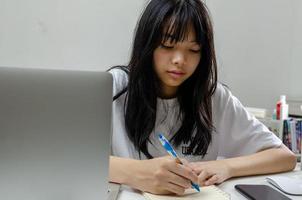 niña sosteniendo un bolígrafo escribiendo un libro de estudio aprendiendo un video digital de internet en línea computadora portátil en casa. foto