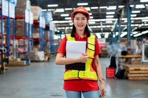 Smart smiling Asian woman working in store warehouse. She is standing and holding document folder and writing some note. She is feeling happy to work. Smart working girl concept. photo