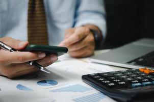 Businessman hand holding mobile phone and pen with graphs and charts business documents on desk. Calculator and computer for investment and economic analysis. Financial and tax business concept. photo