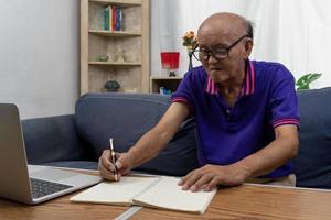 anciano asiático sosteniendo un bolígrafo, escriba una nota de mensaje y una computadora portátil en la mesa en el sofá. foto