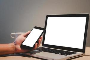 man hand holding mobile smartphone and computer laptop mock up blank screen on desk. photo