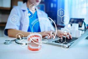 dentista concentrado sentado en la mesa con muestras de mandíbula modelo de diente y trabajando con tableta y computadora portátil en la clínica dental profesional de la oficina dental. con holograma de icono virtual foto