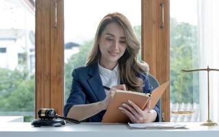 joven abogada asiática confiada y exitosa o consultora legal de negocios leyendo un libro de leyes o escribiendo algo en su cuaderno en el escritorio de su oficina. foto