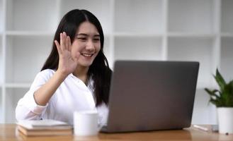 Pretty and charming young Asian female employee having a video call with her team via laptop computer. Remote working concept photo