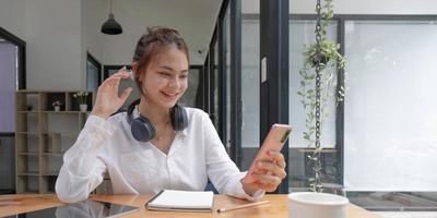 feliz joven adolescente asiática sentada en un sofá en casa sosteniendo el teléfono mirando la pantalla agitando la mano video llamando a un amigo a distancia en línea en la aplicación de chat móvil usando la aplicación de videochat de smartphone. foto