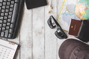 Top view of Keyboard with calendar,smart phone,earphones,sunglasses,cap,wallet,passport,map and globe on white wooden table background, Travel concept photo