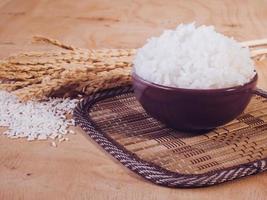 arroz cocido en tazón con grano de arroz crudo y planta de arroz seco sobre fondo de mesa de madera. foto