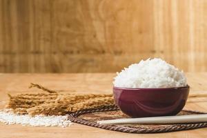 arroz cocido en tazón con grano de arroz crudo y planta de arroz seco sobre fondo de mesa de madera. foto