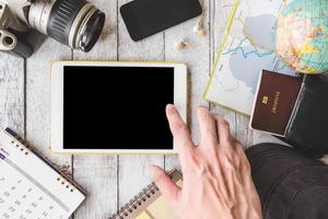 Top view of Male hands using tablet with camera,calendar,smart phone,earphones,cap,notebook,wallet,passport,map and globe on white wooden table background, Travel concept photo