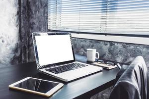 portátil con pantalla blanca en blanco, taza blanca de café, tableta, portátil, bolígrafo y gafas en el escritorio foto