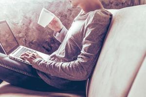 Casual young man using laptop and holding a cup of coffee on sofa at home. photo