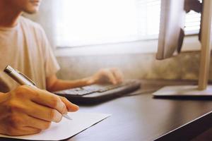 Casual young man writing some data in paper and working on computer, sitting at desk, toned with sunlight photo