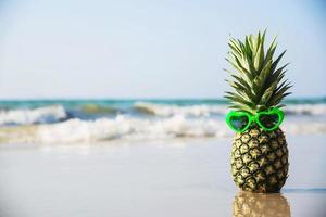 Encantadora piña fresca poner gafas de sol en forma de corazón en la playa de arena limpia con fondo de olas marinas - fruta fresca con concepto de fondo de vacaciones de sol de arena de mar foto