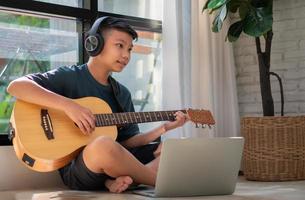 Asian boy playing guitar and watching online course on laptop while practicing for learning music or musical instrument online at home. Boy students study online with video call teachers play guitar. photo