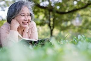 feliz anciana asiática y usa un reloj de salud y un libro de lectura y se acuesta en la alfombra de picnic en el parque y una canasta de frutas además. concepto de anciana feliz después de la jubilación y buena salud foto