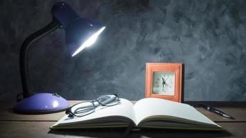 Lamp with notebook,pen,clock and glasses  on  wooden table with vintage wall background. photo