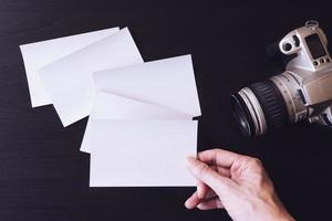 vista superior de la tarjeta fotográfica en blanco en la mano con la cámara en el escritorio de la oficina. foto