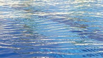 El material de archivo de fondo de la superficie del agua presenta un primer plano extremo del agua azul en una piscina. el agua se ondula en cámara lenta y tiene algunos reflejos del sol. video