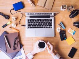 Top view of Male hands holding a coffee cup, Laptop with office supplies on wooden desk. photo