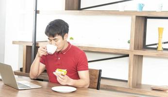 un joven asiático sonriente con una camiseta roja casual disfruta desayunando, comiendo un sándwich, un joven cocinando comida y bebiendo en la cocina estilo loft foto