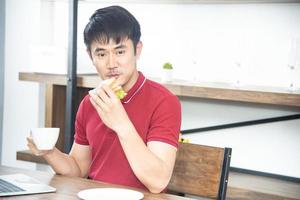 un joven asiático sonriente con una camiseta roja casual disfruta desayunando, comiendo un sándwich, un joven cocinando comida y bebiendo en la cocina estilo loft foto