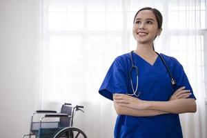 Young Asian nurse smiling happily with a hand on hip and confident, positive, proud and friendly attitude against white curtain with wheelchair in background photo