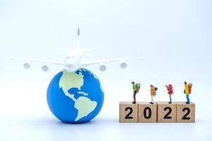 2022 New Year and Travel Concept. Closeup of group of traveler miniature figures with backpack standing on stack of wooden number block with airplane model on mini world ball on white background. photo