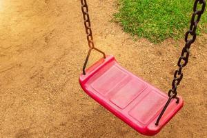 A red swing is attached to two old rusted chains in the playground. in the park saw the ground and the grass photo