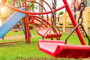 A red swing is attached to two old rusted chains in the playground. in the park saw the ground and the grass photo