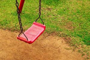 un columpio rojo está unido a una vieja cadena oxidada en el patio de recreo. en el parque vio el suelo y la hierba foto