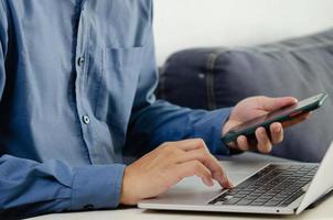 mano de hombre de negocios escribiendo teclado en computadora portátil con teléfono inteligente. cierra a un hombre de negocios que comunica internet y las redes sociales en su escritorio. foto