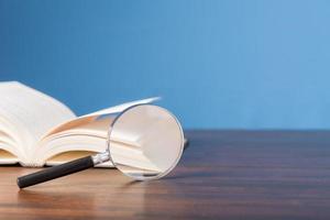 open book with magnifying glass on wooden desk in information library of school or university, concept for education,reading , study, copy space and blue background. photo