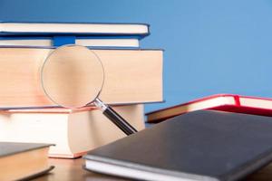 Stack book with magnifying glass on wooden desk in information library of school or university, concept for education, reading, study, copy space on blue background. photo